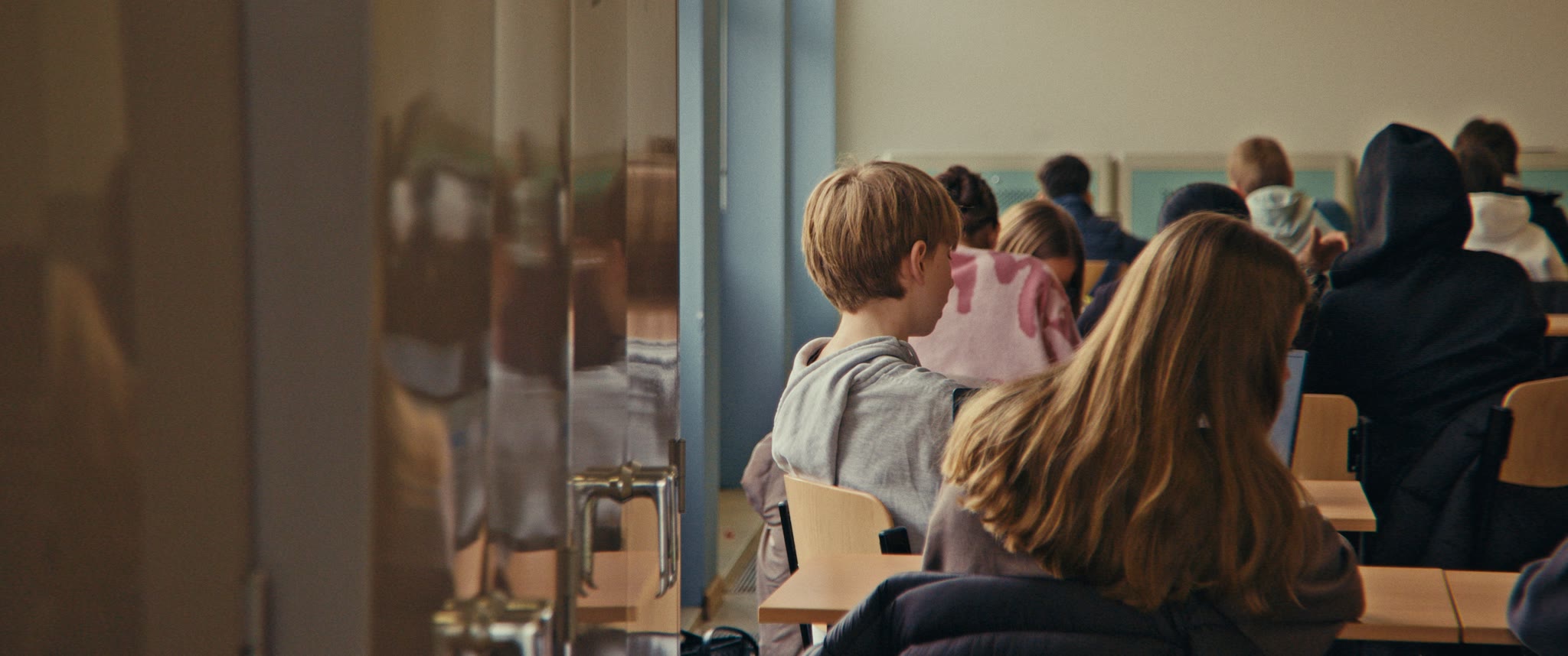 Een klas gezien van achteren: leerlingen aan hun banken, een jongen kijkt opzij.